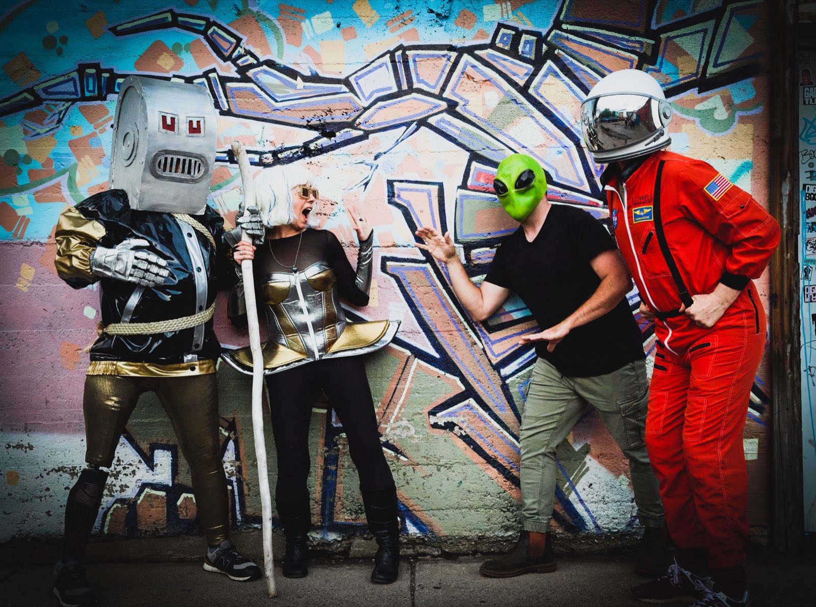 Box-headed robot, metallic performers, alien, and astronaut posing before graffiti