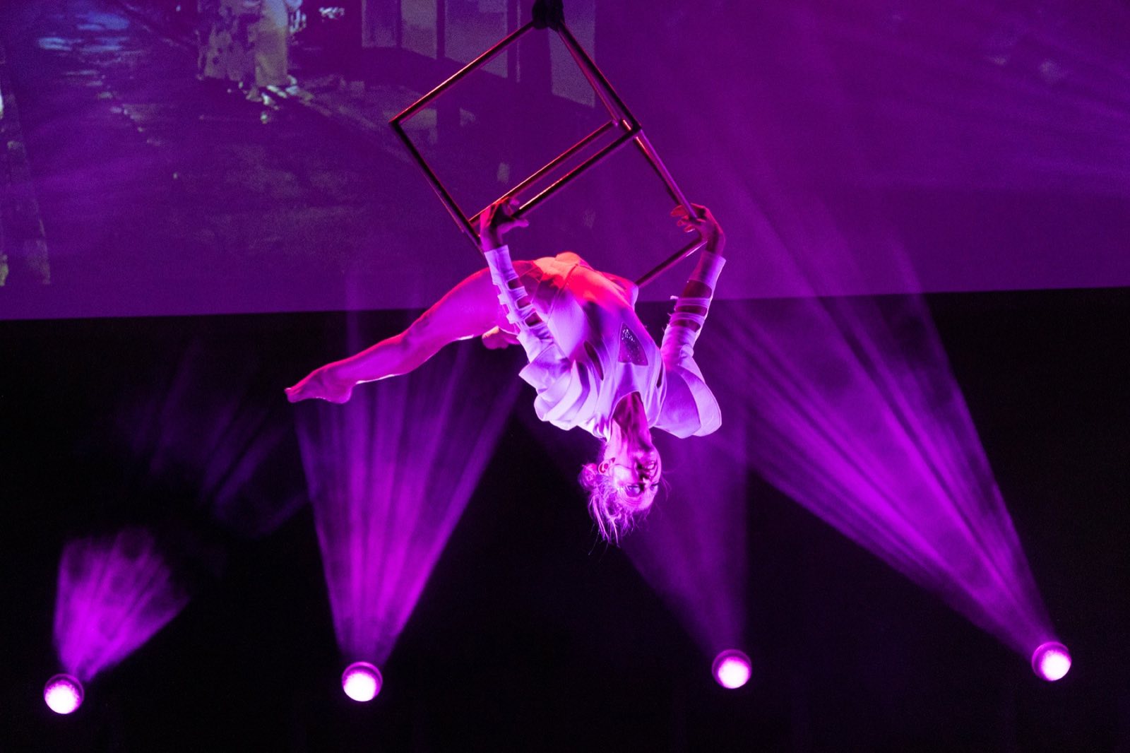 Aerialist hanging beneath a metal cube in magenta light