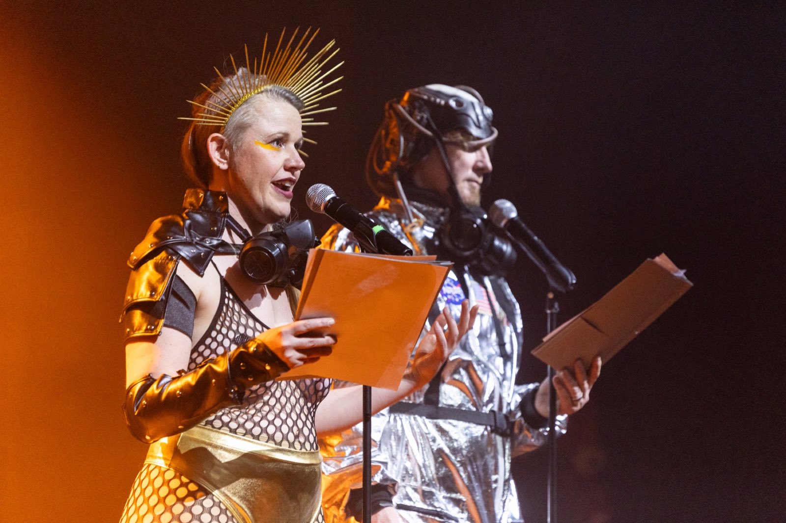 Two costumed narrators reading from clipboards under warm stage light