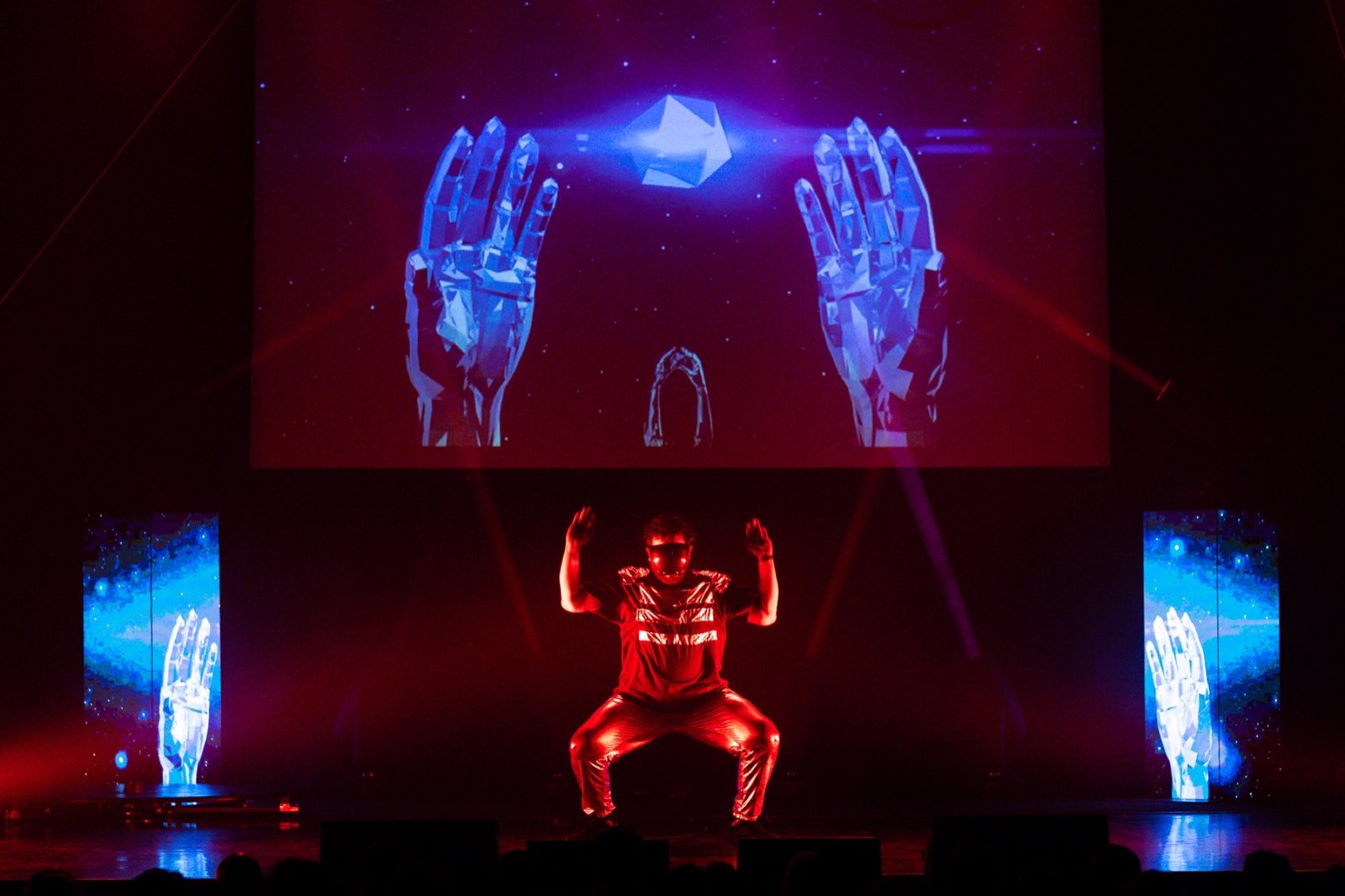 Solo performer kneeling beneath a projection of enormous robotic hands