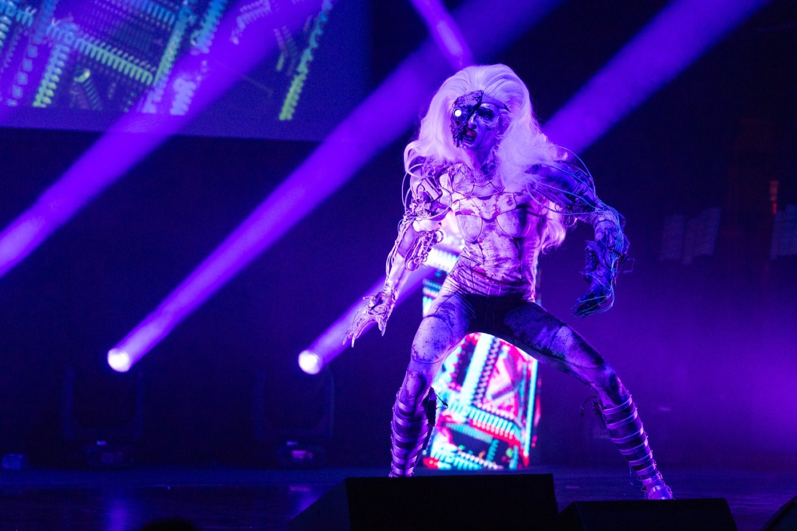 White-haired costumed character standing in crossing purple spotlights