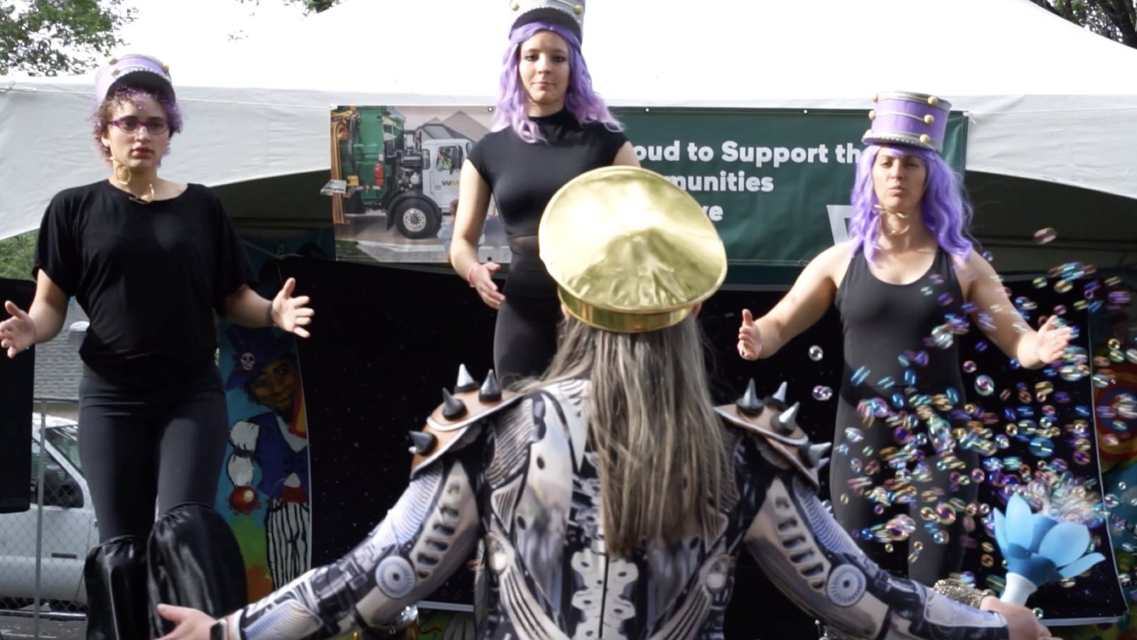 Crowned robot character facing three black-clad performers on the festival stage
